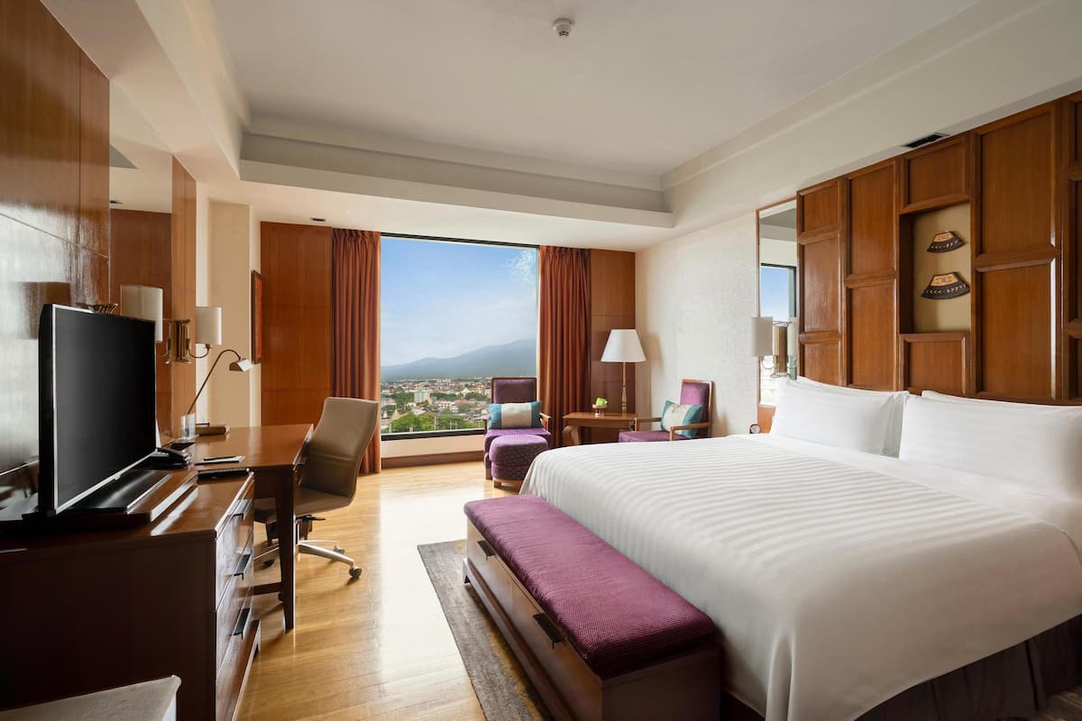 A hotel room with a large bed, a desk with a TV, a chair, a desk lamp, and a view of the city.