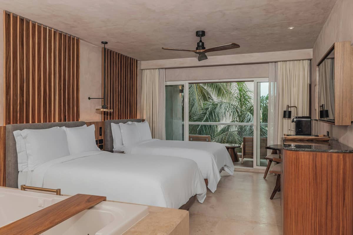 A modern hotel room with two beds, a wooden accent wall, a ceiling fan, and a view of palm trees through large windows.