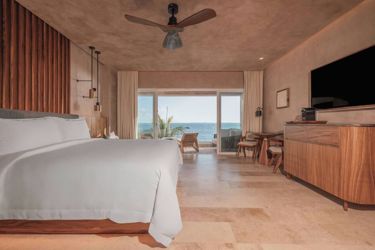 A hotel room with a large bed, a ceiling fan, a TV, a wooden dresser, and a view of the sea.