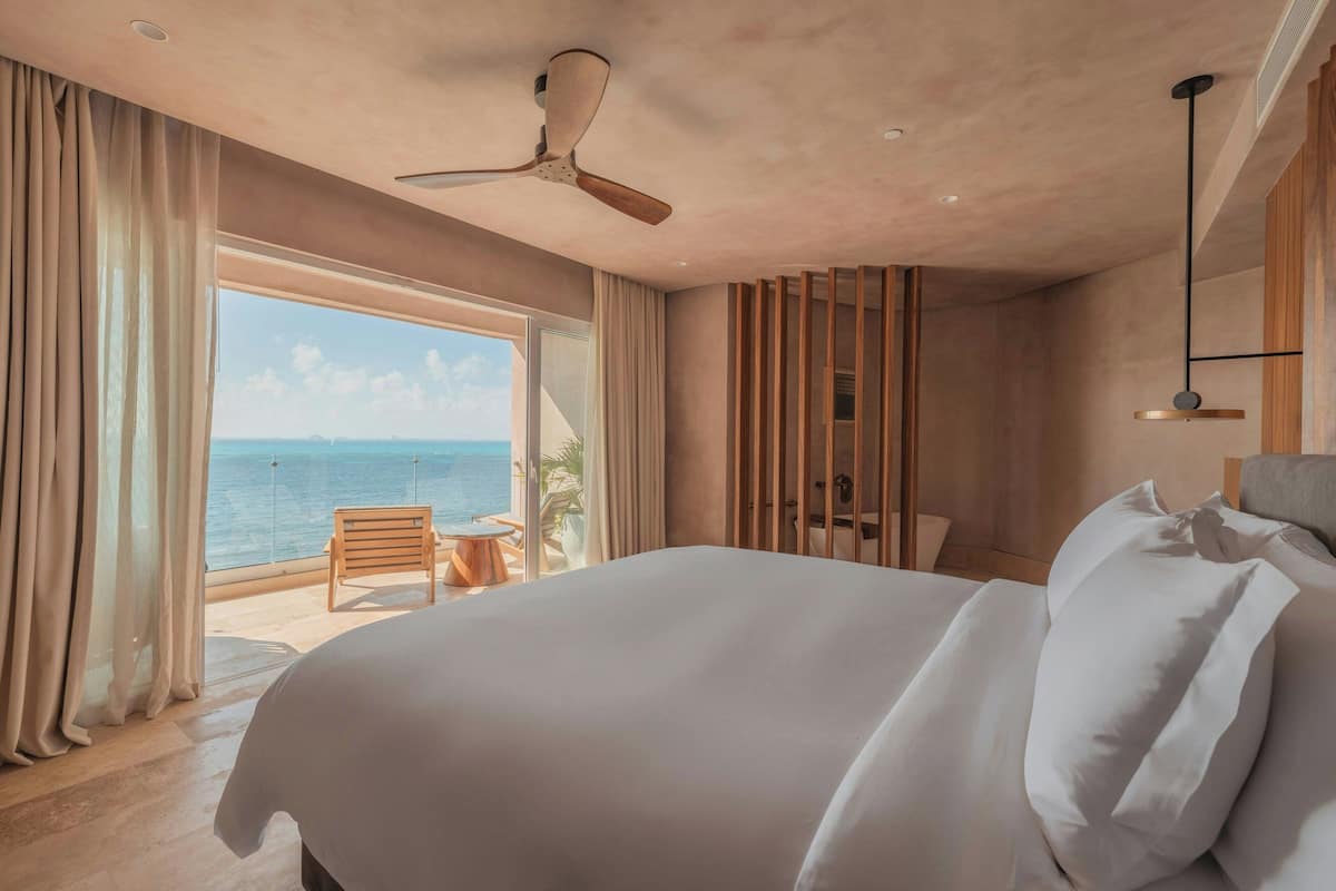 A hotel room with a large bed, a ceiling fan, a view of the ocean, and a balcony with a table and chair.