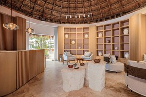 A spacious lobby with a thatched ceiling, wooden paneling, and a central seating area with two round tables and chairs.