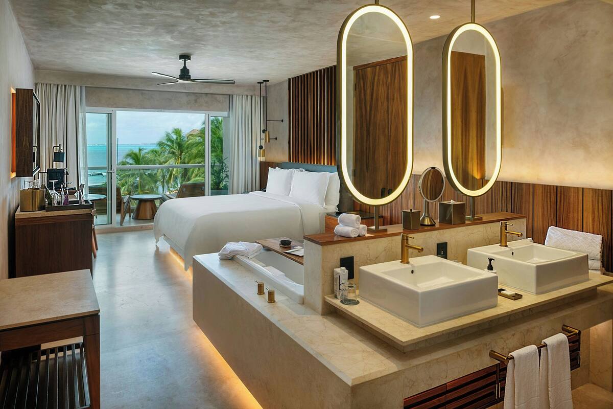 A modern hotel room with a large bed, two sinks, a vanity, and a balcony with a view of the ocean.