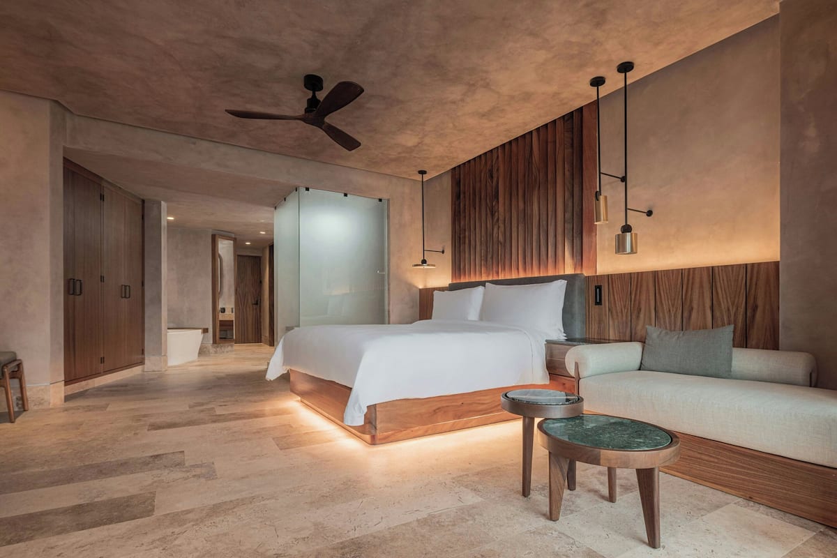 A modern hotel room with a large bed, a sofa, a round table, and a ceiling fan.