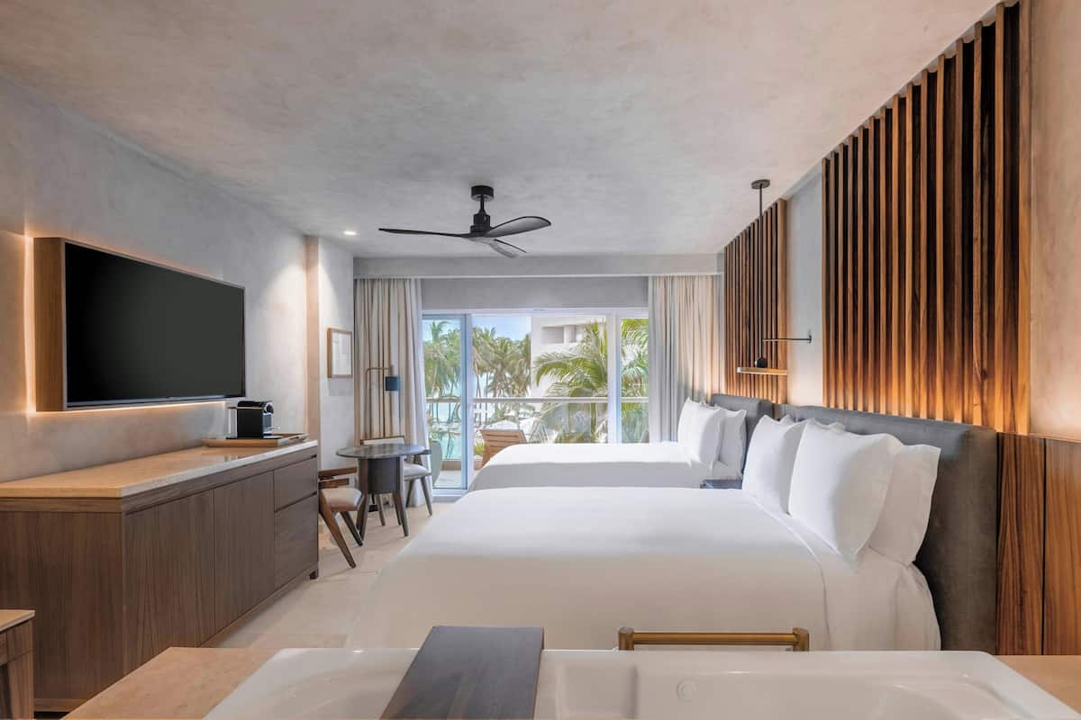 A modern hotel room with a large bed, a flat-screen TV, a wooden cabinet, a desk, and a balcony with a view of palm trees.