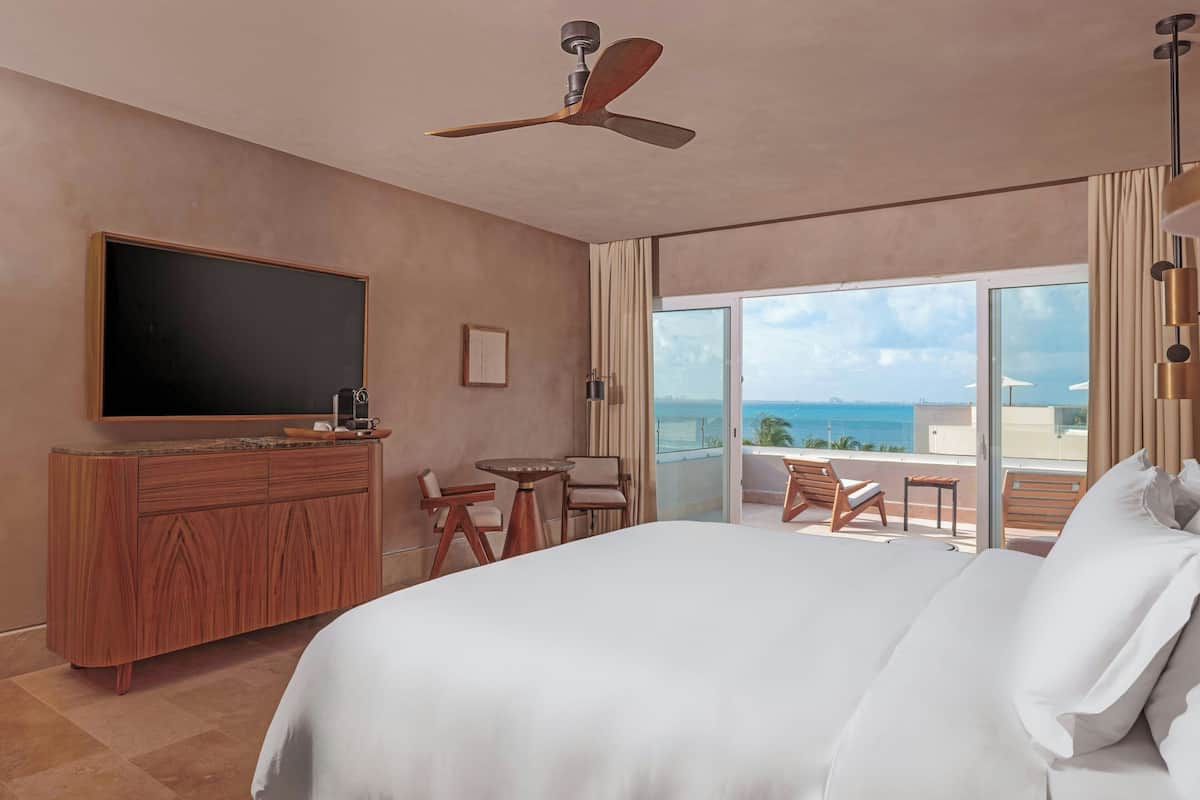 A hotel room with a bed, a TV, a desk, and a balcony with a view of the ocean.