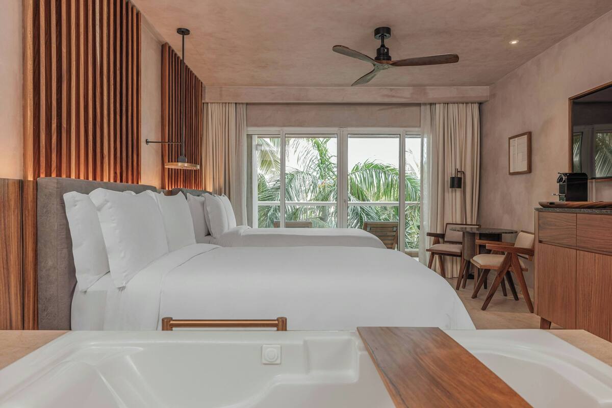 A hotel room with a large bed, a desk, a chair, a coffee machine, and a view of palm trees.