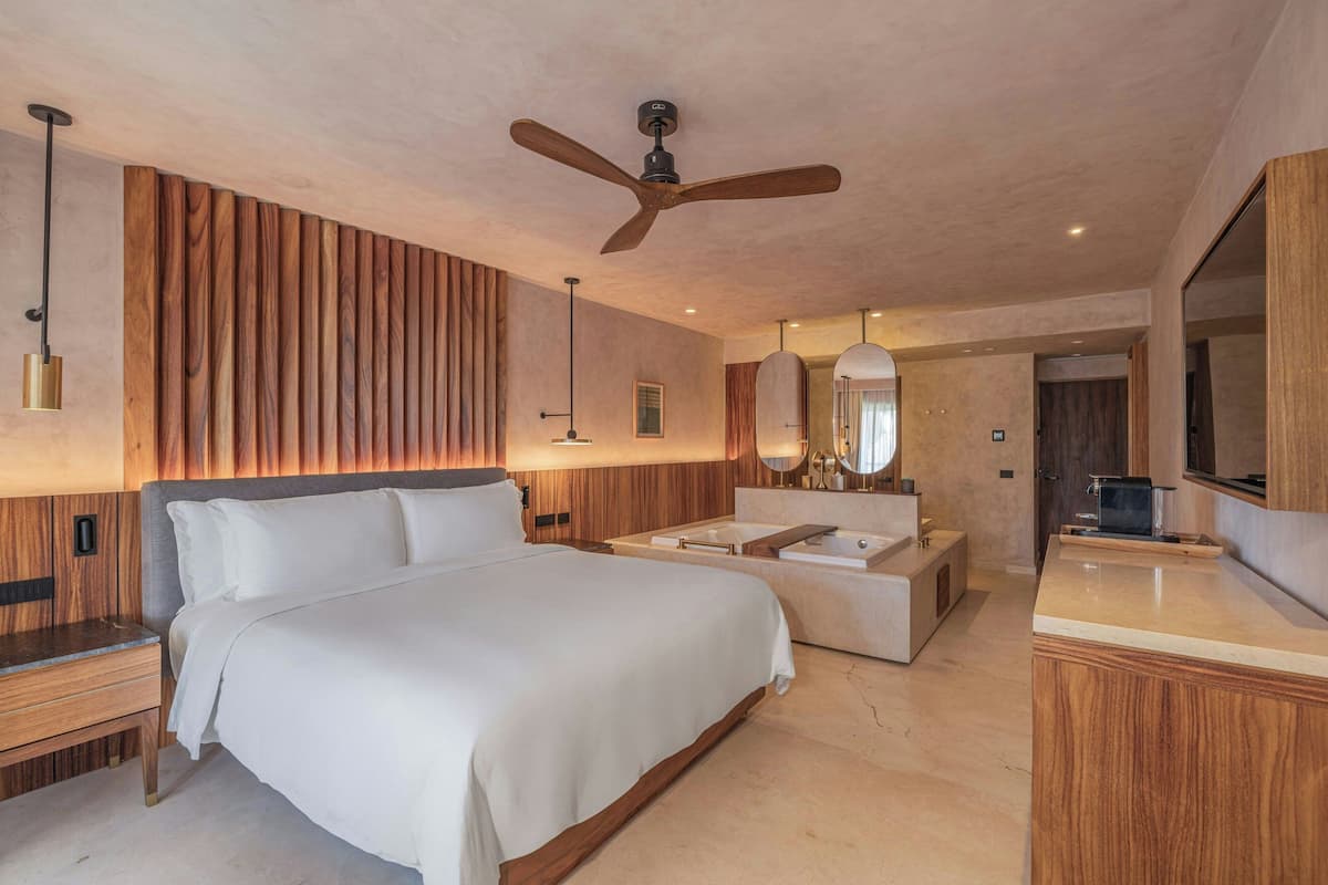 A modern hotel room with a large bed, a vanity with a bathtub, and a ceiling fan.