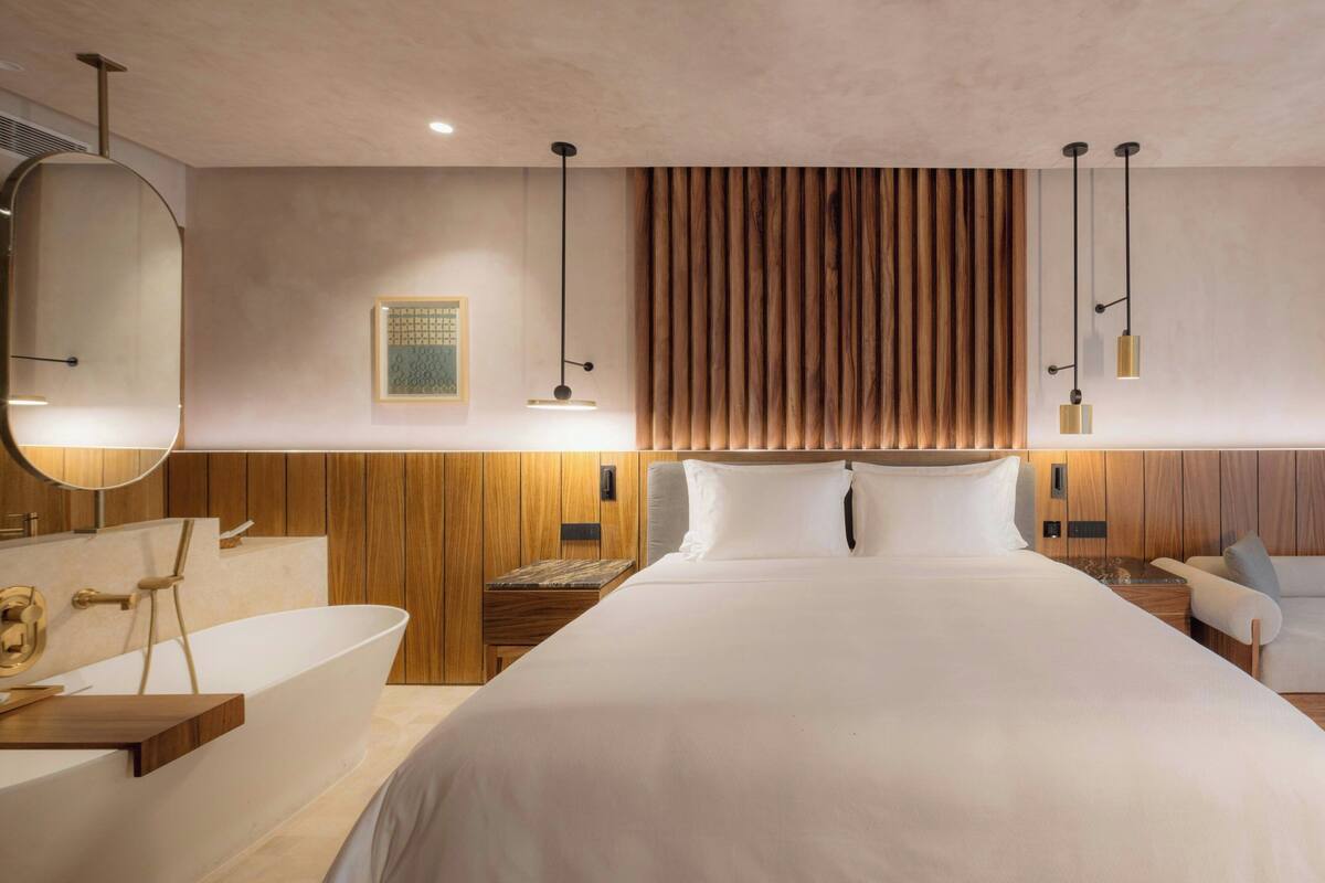 A modern hotel room with a large bed, a bathtub, a sitting area, and a wooden panel wall.