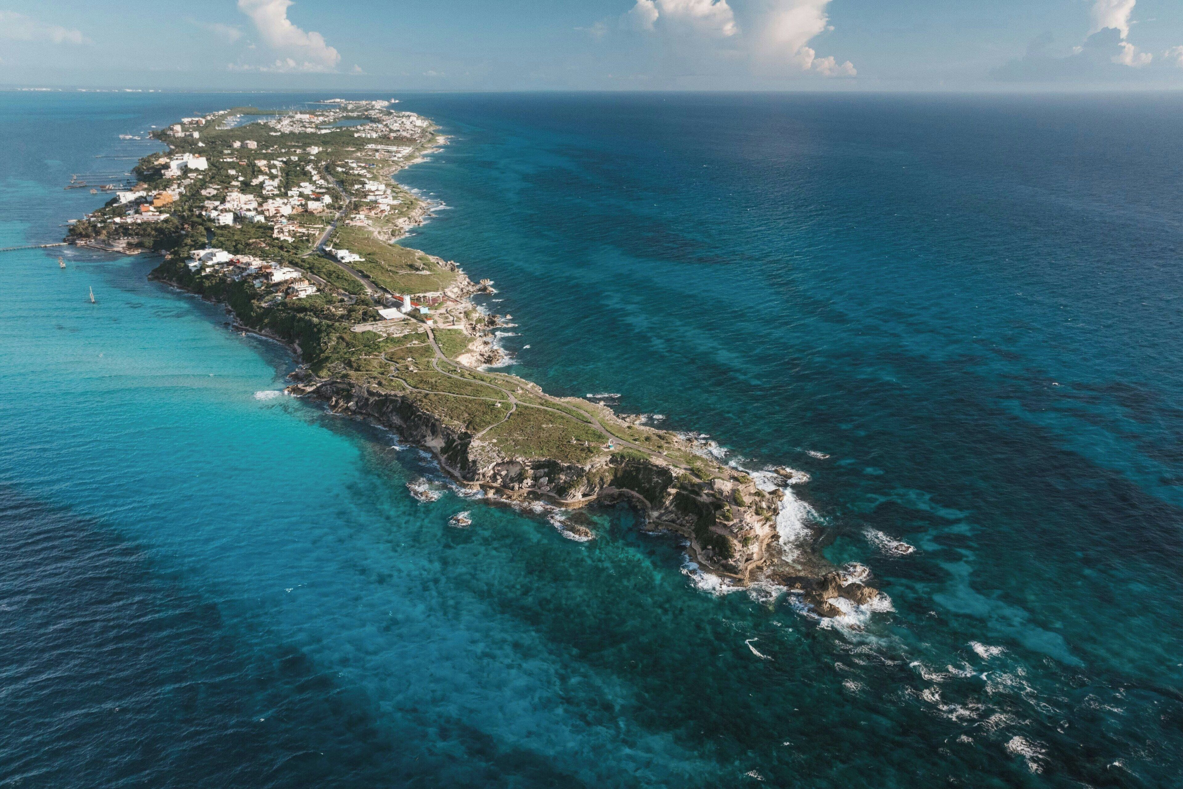 An aerial view of a coastal island with a narrow strip of buildings along the shoreline, surrounded by clear blue waters.