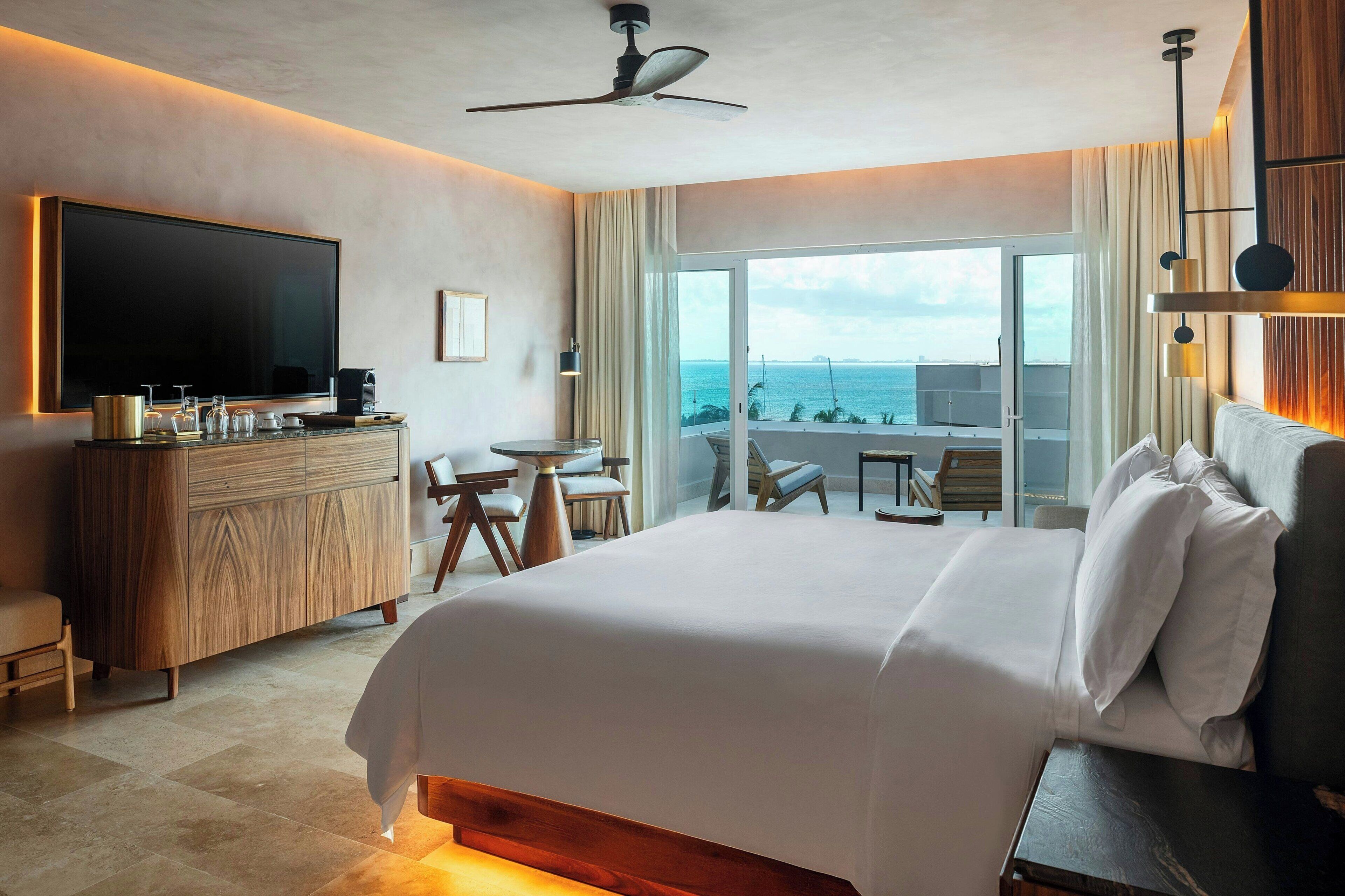 A hotel room with a large bed, a TV, a desk, and a balcony with a view of the sea.