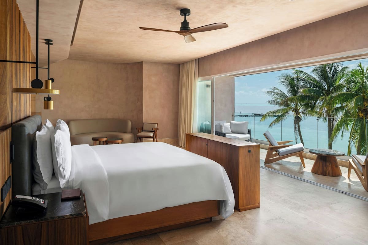 A modern hotel room with a large bed, a desk, a chair, and a balcony with a view of the ocean and palm trees.