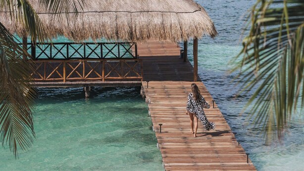 Une jetée en bois mène à un bungalow sur pilotis doté d’un toit de chaume, surplombant une eau turquoise cristalline et offrant une vue lointaine sur la ville.