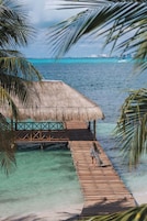 Un muelle de madera conduce a una cabaña sobre el agua con techo de paja, con agua turquesa cristalina y un horizonte urbano distante.