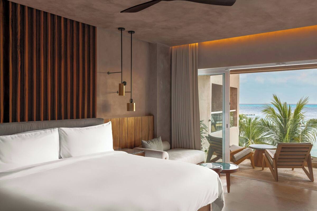 A hotel room with a large bed, a small seating area with a table, and a balcony with a view of the ocean.