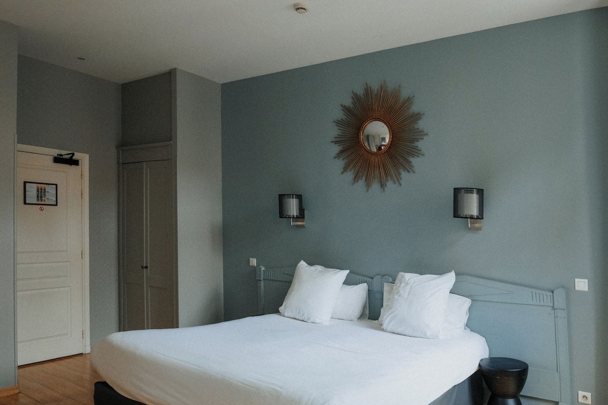 A bedroom with a large bed, a bedside table, a wall-mounted mirror, and a door.