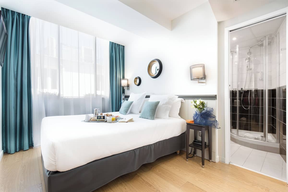 A hotel room with a large bed, a bedside table with a lamp, a shower area, and a view of the city.