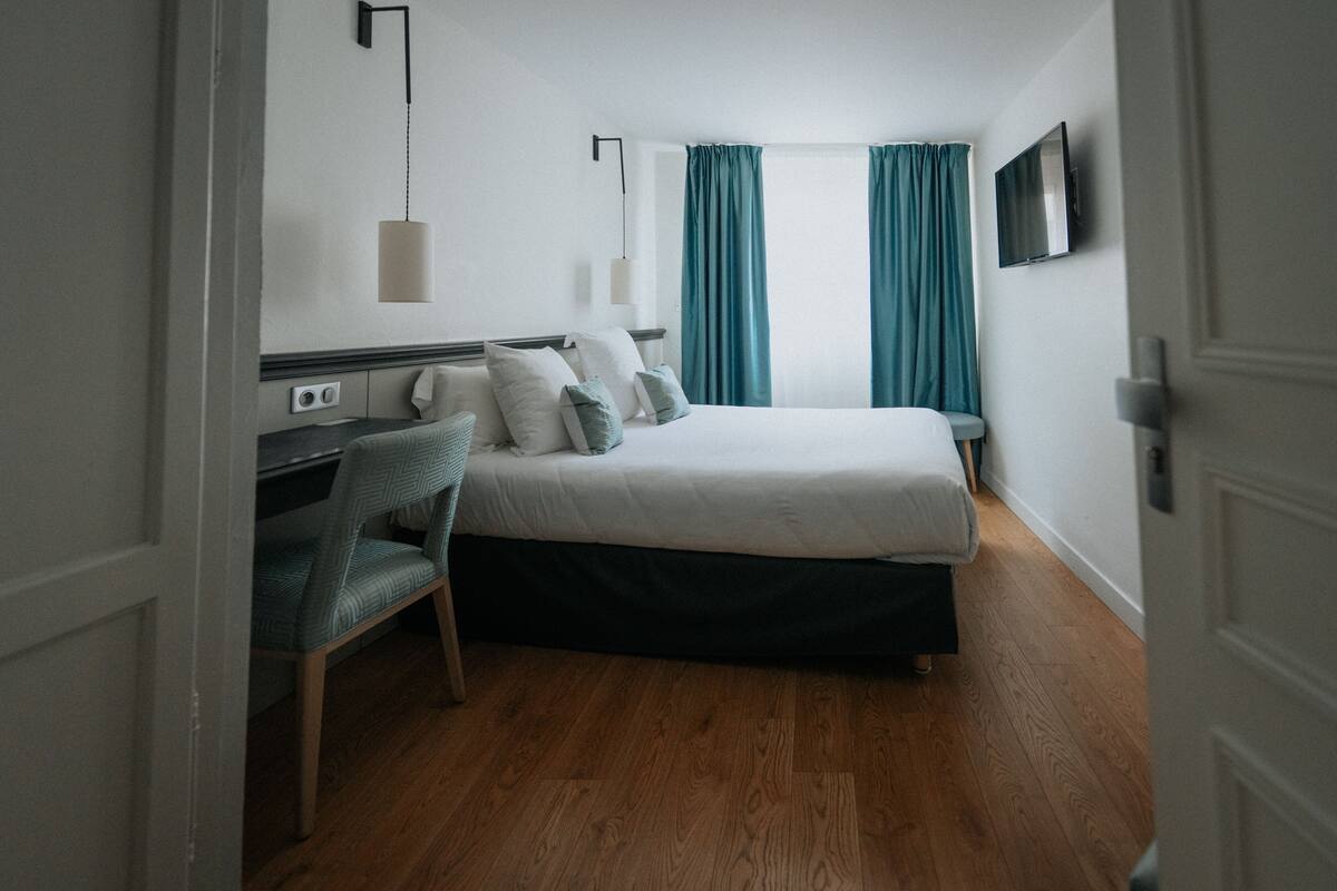 A hotel room with a bed, a desk, a chair, a TV, and a window with curtains.