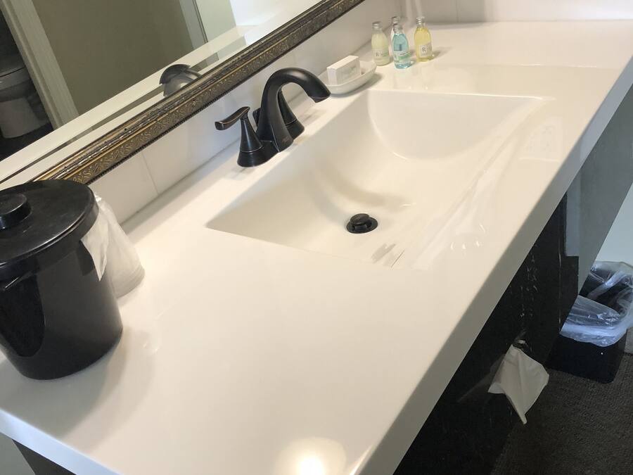 Ein Badezimmer mit einem weißen Waschbecken, einem schwarzen Wasserhahn und einem Spiegel darüber. Auf dem Waschbecken stehen Toilettenartikel und darunter befindet sich ein Mülleimer.