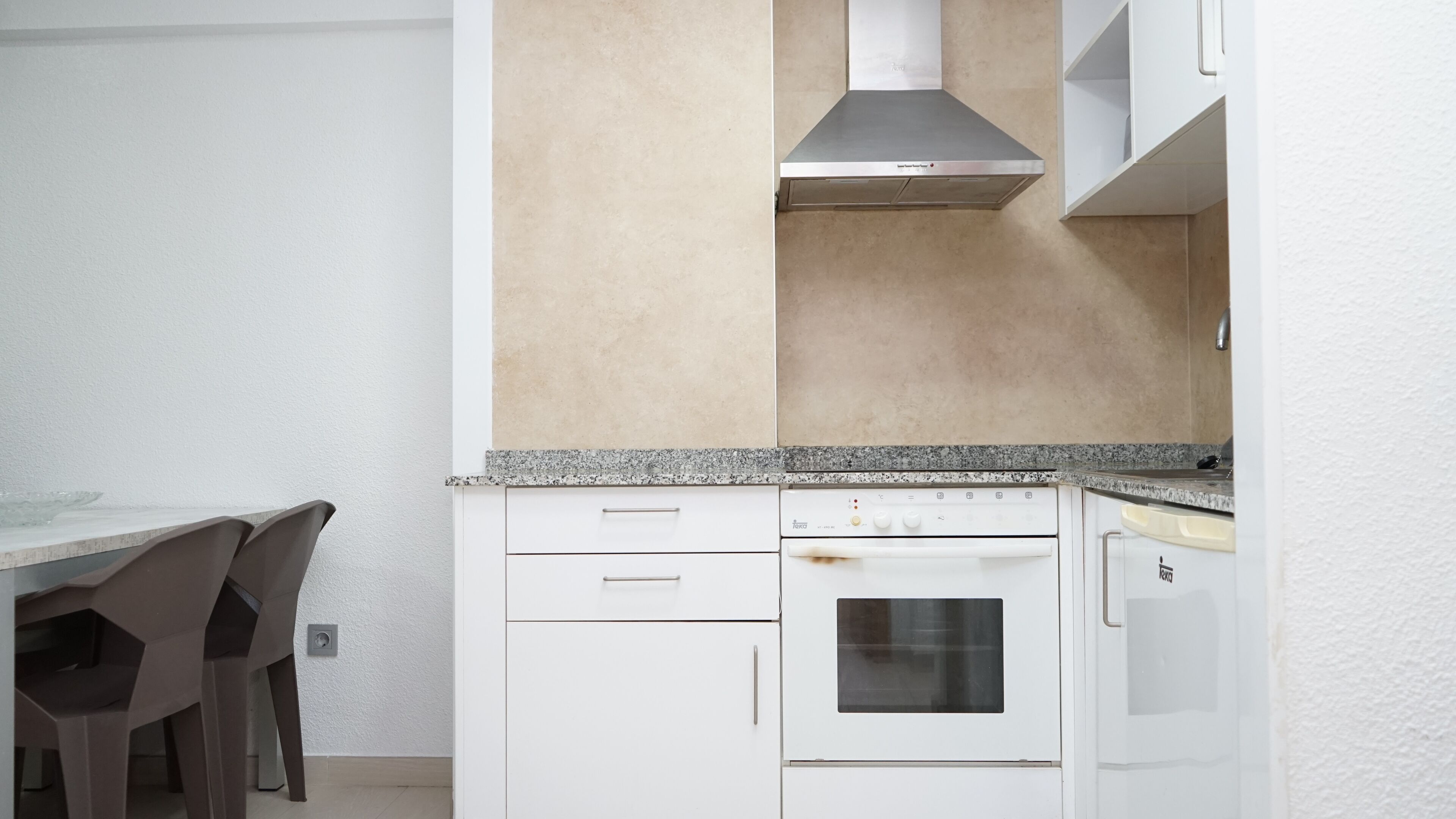 A compact kitchen with white cabinets, a stainless steel range hood, a built-in oven, and a dishwasher. There is a granite countertop and a dining area with a table and chairs.