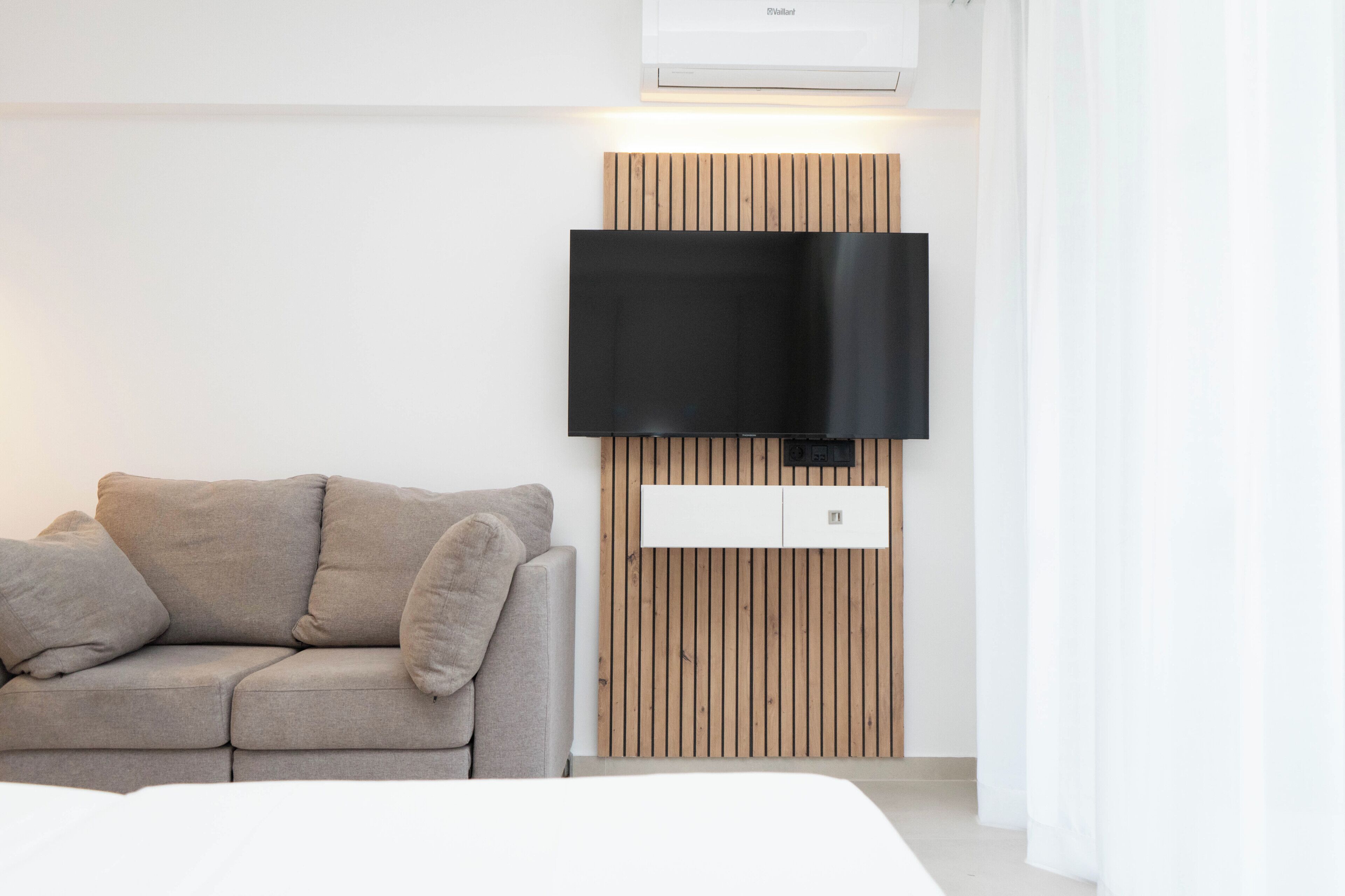A modern room with a flat-screen TV mounted on a wooden panel wall, a grey sofa, and a white bed.