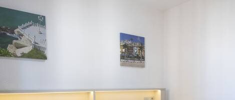 Una cama bien hecha con sábanas blancas y un plaid gris, un cabecero con iluminación integrada y dos cuadros colgados en la pared que representan escenas costeras.