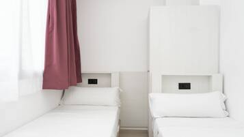 Una habitación pequeña y limpia con dos camas individuales, una ventana con cortinas translúcidas y una pared blanca.
