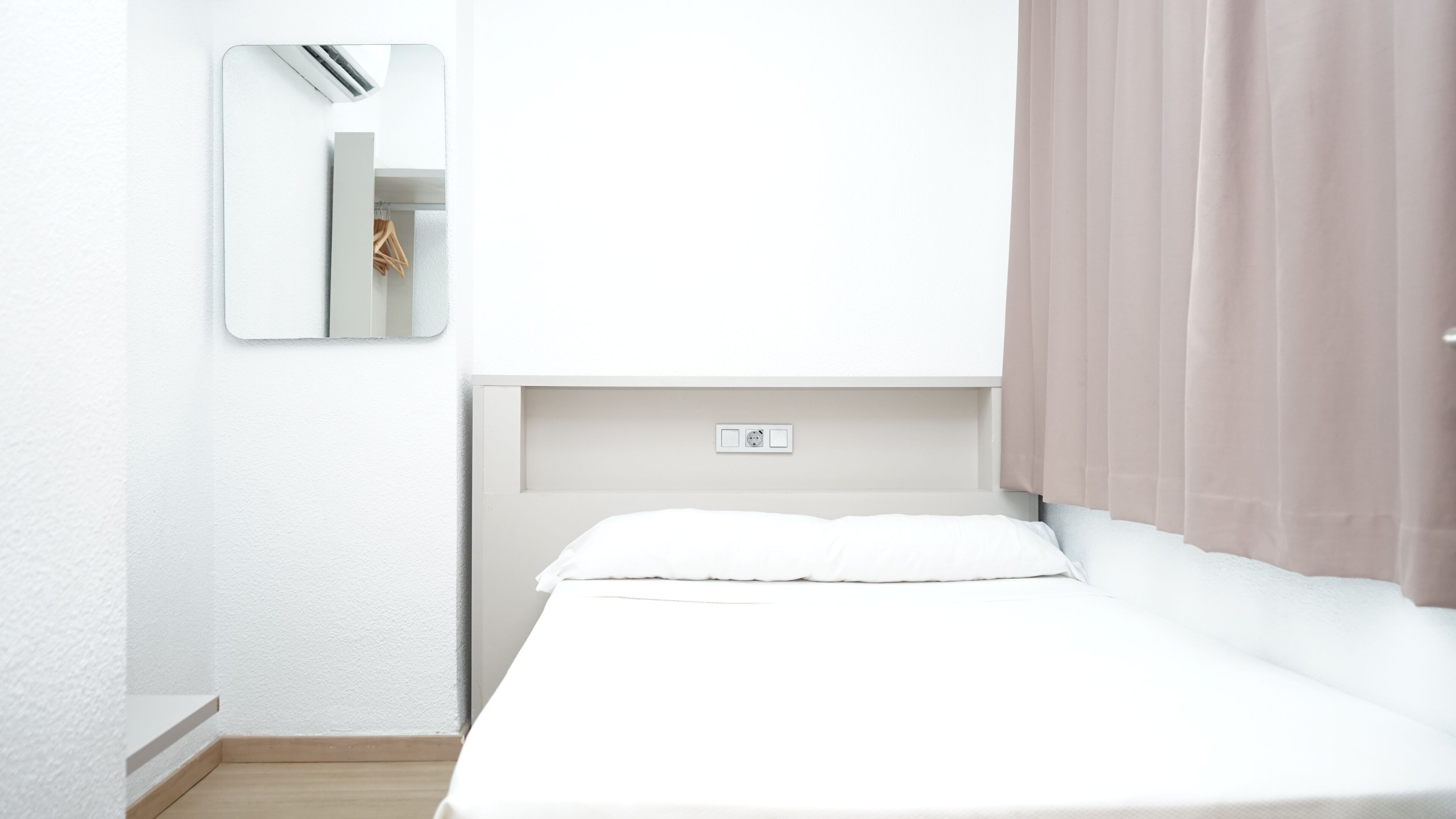 Minimalistinen hotellihuone, jossa on yksi sänky, seinään asennettu ilmastointilaite, peili ja ikkuna, jossa on verhot.