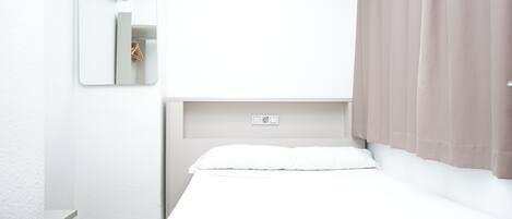 Minimalistinen hotellihuone, jossa on yksi sänky, seinään asennettu ilmastointilaite, peili ja ikkuna, jossa on verhot.