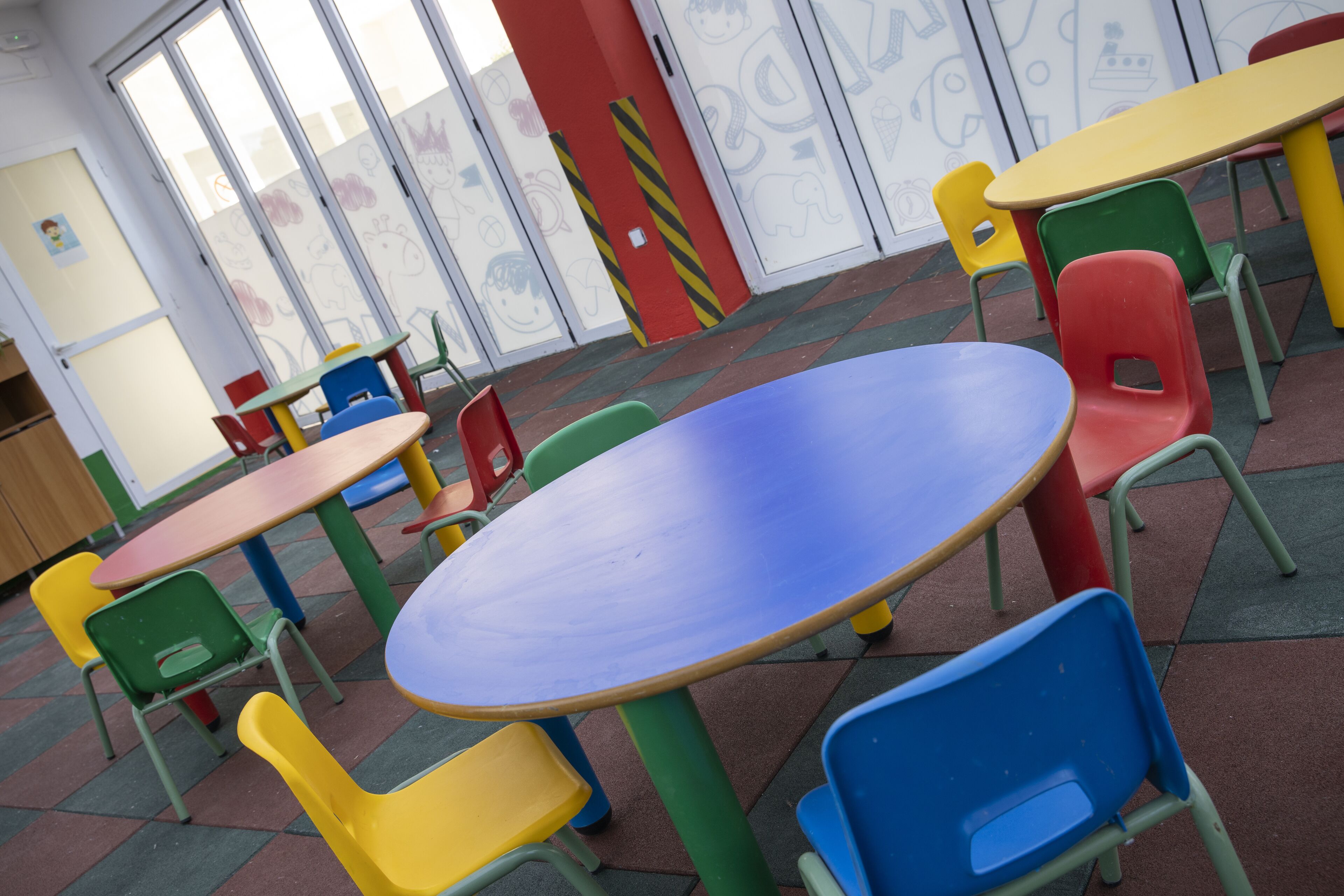 A children's play area with colorful tables and chairs, a red door, and a wall with children's drawings.