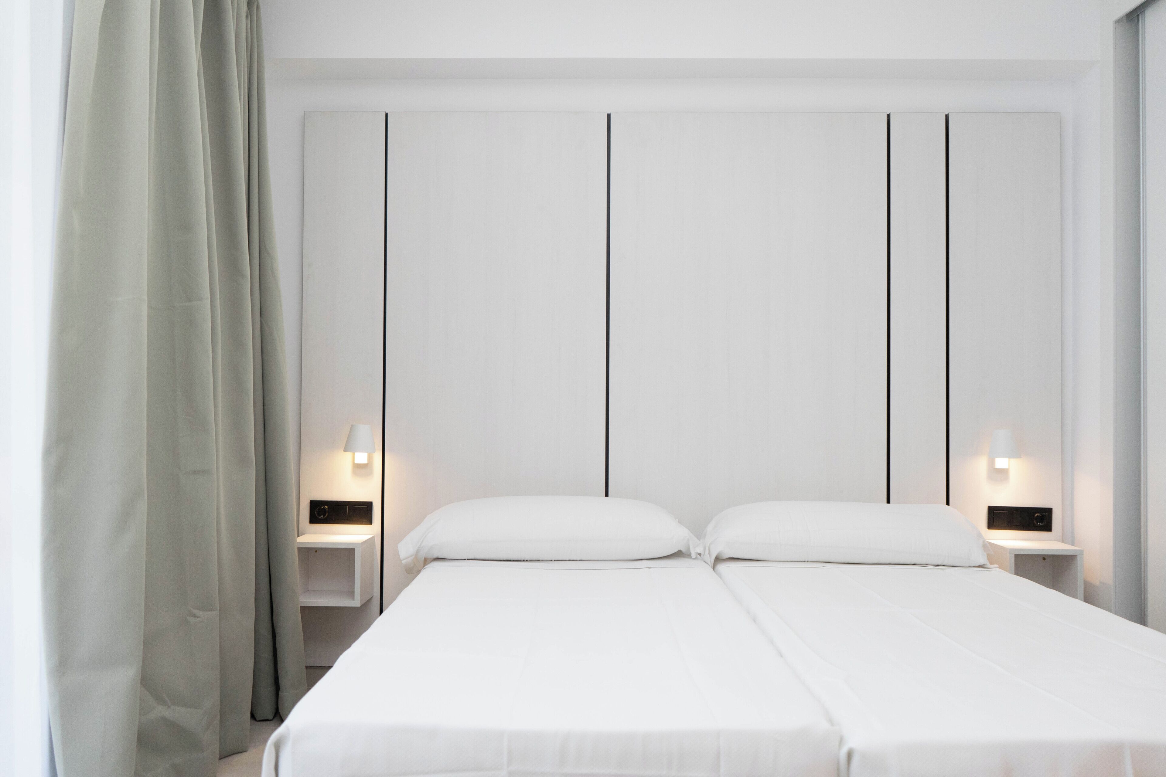 A double bed with white bedding, two bedside tables with lamps, and a plain wall with vertical lines.