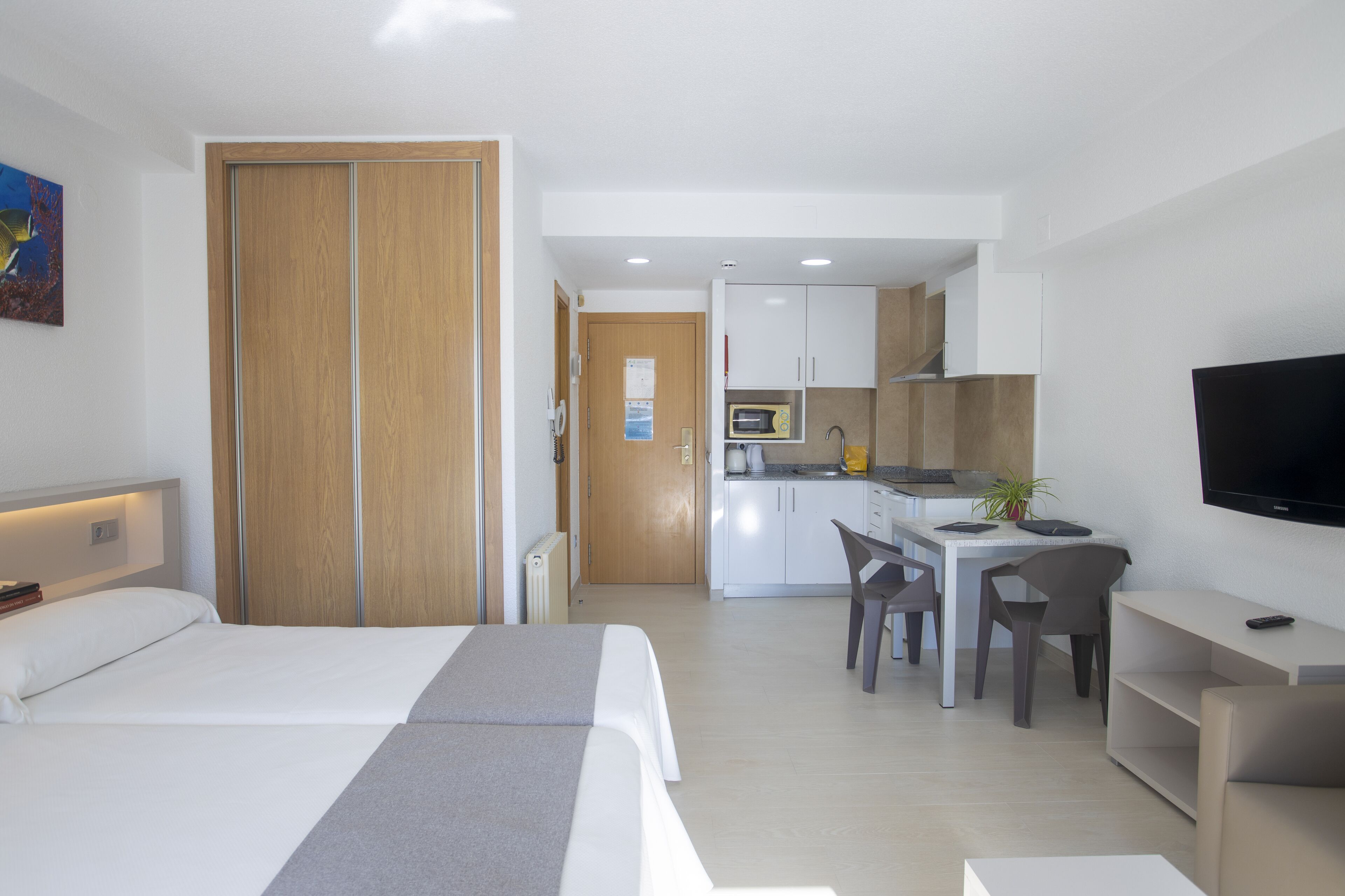 A modern hotel room with a bed, a desk, a chair, a TV, and a kitchenette.