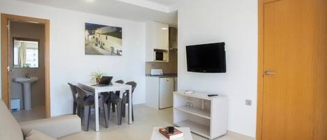 A modern living room with a sofa, a dining table with chairs, a TV mounted on the wall, and a kitchen area in the background.