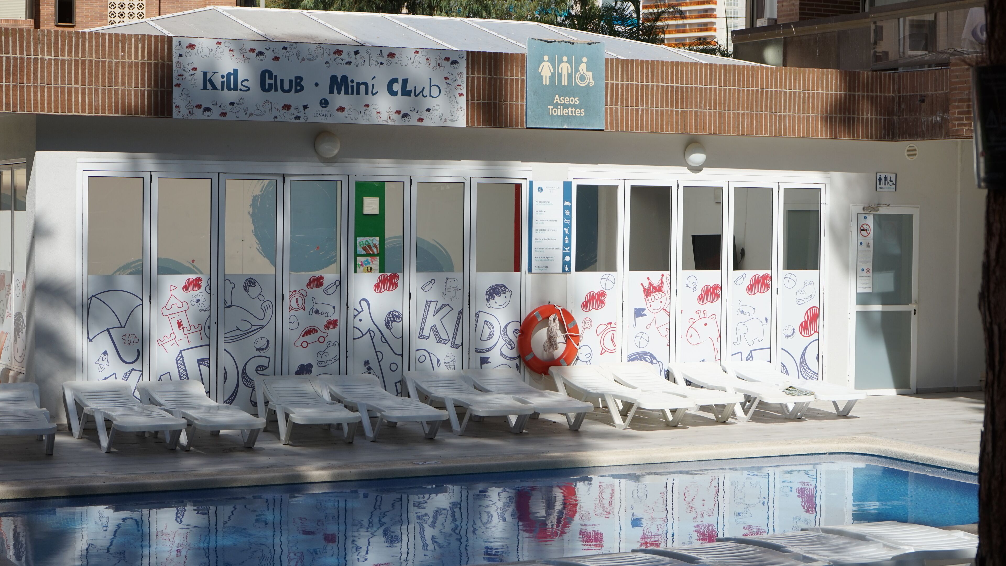 A poolside area with lounge chairs, a lifebuoy, and a sign indicating 'Aseos Toilettes' (toilets).