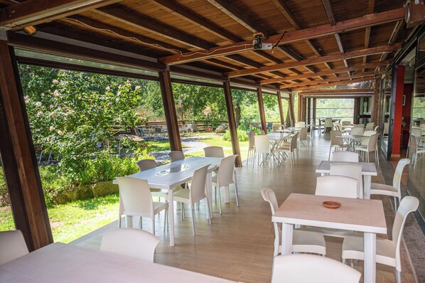 Un área de comedor al aire libre con mesas y sillas blancas, rodeada por paredes de vidrio y vistas a zonas verdes.