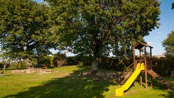 Une aire de jeux avec un toboggan jaune, entourée d’herbe verte et d’arbres.