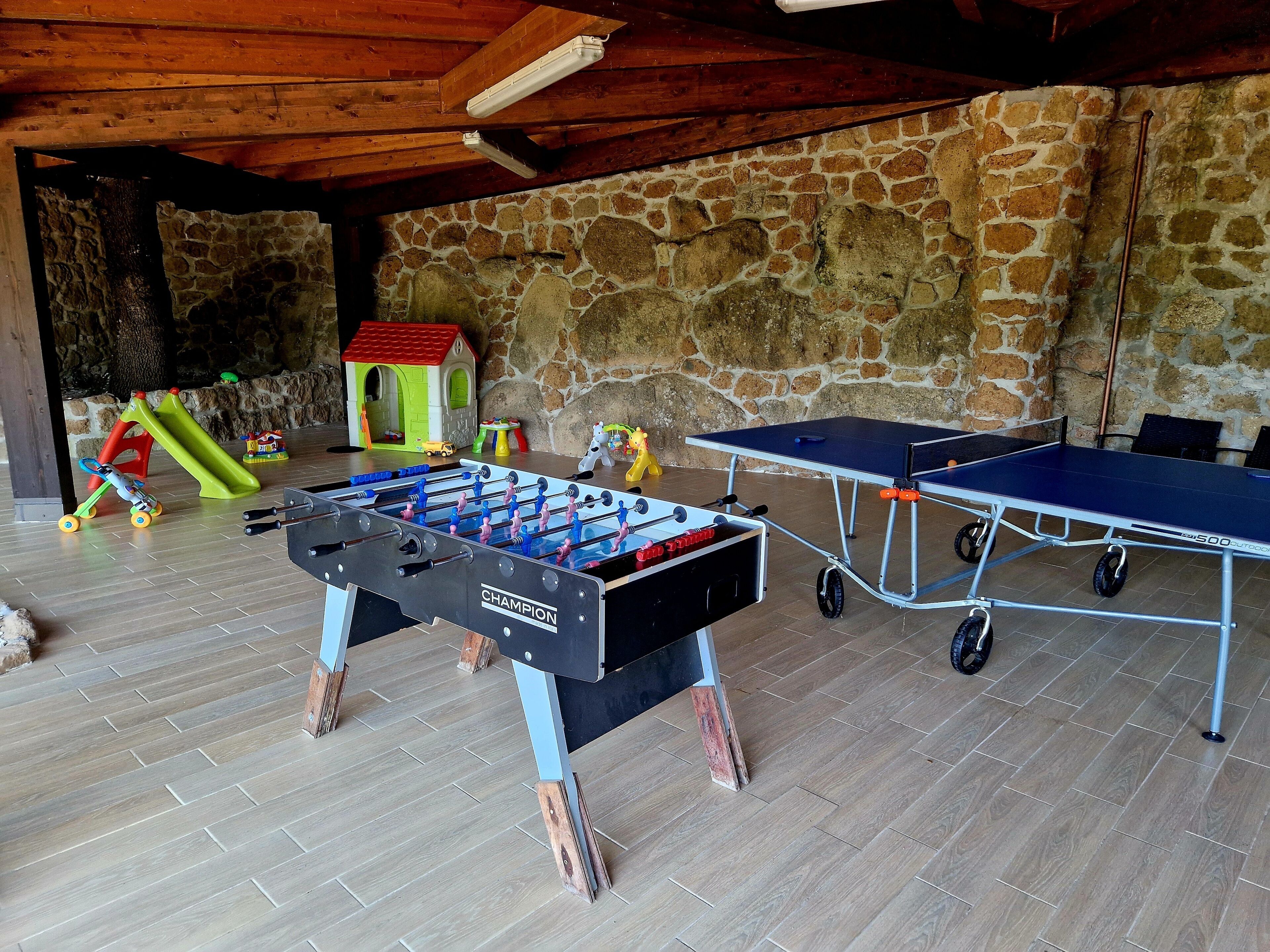 A room with a foosball table, a ping pong table, and a children's play area with a slide and toys.