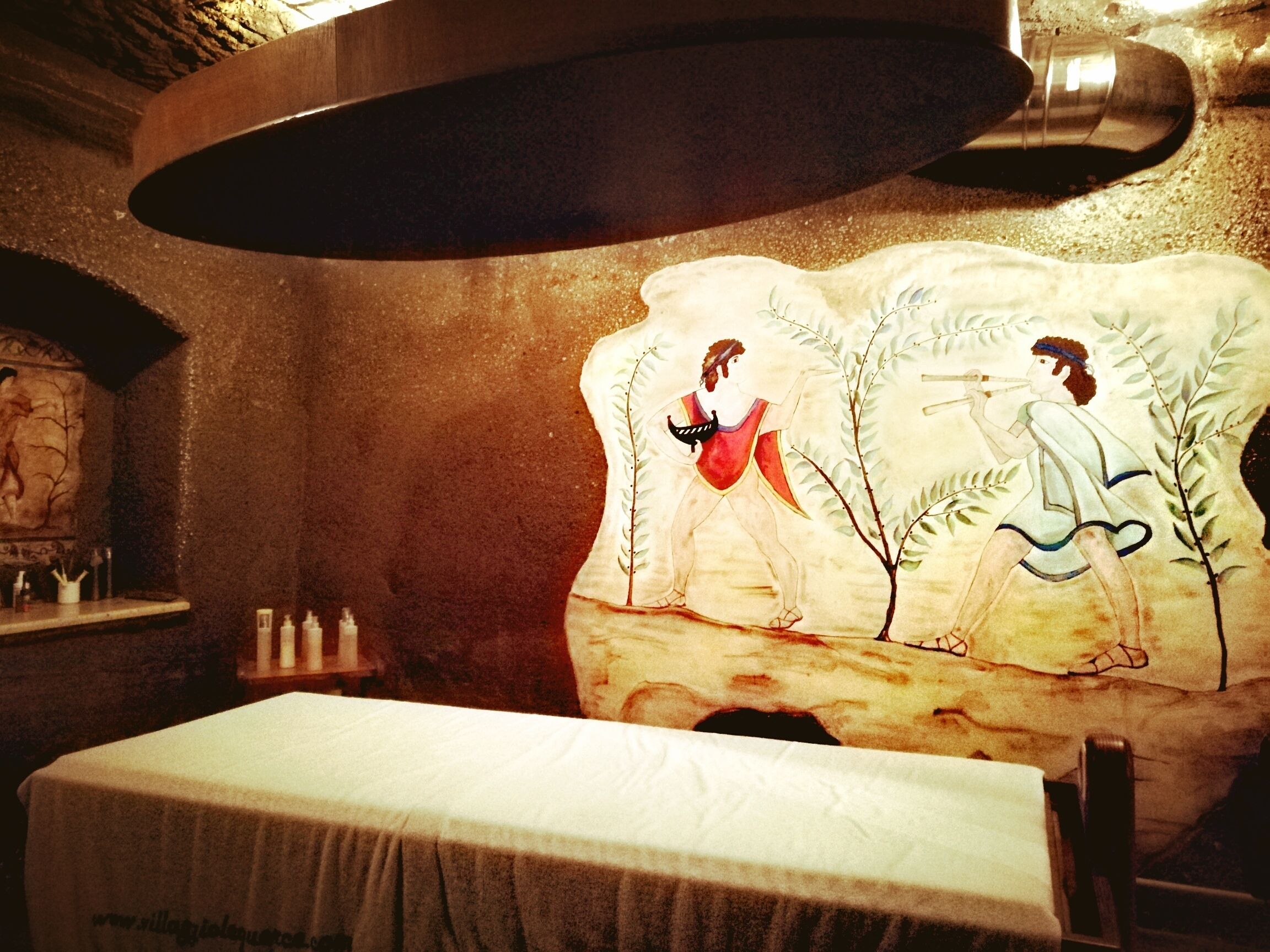Ein Zimmer mit Wandgemälde, das zwei Personen, einen Tisch und ein Regal mit Flaschen zeigt.
