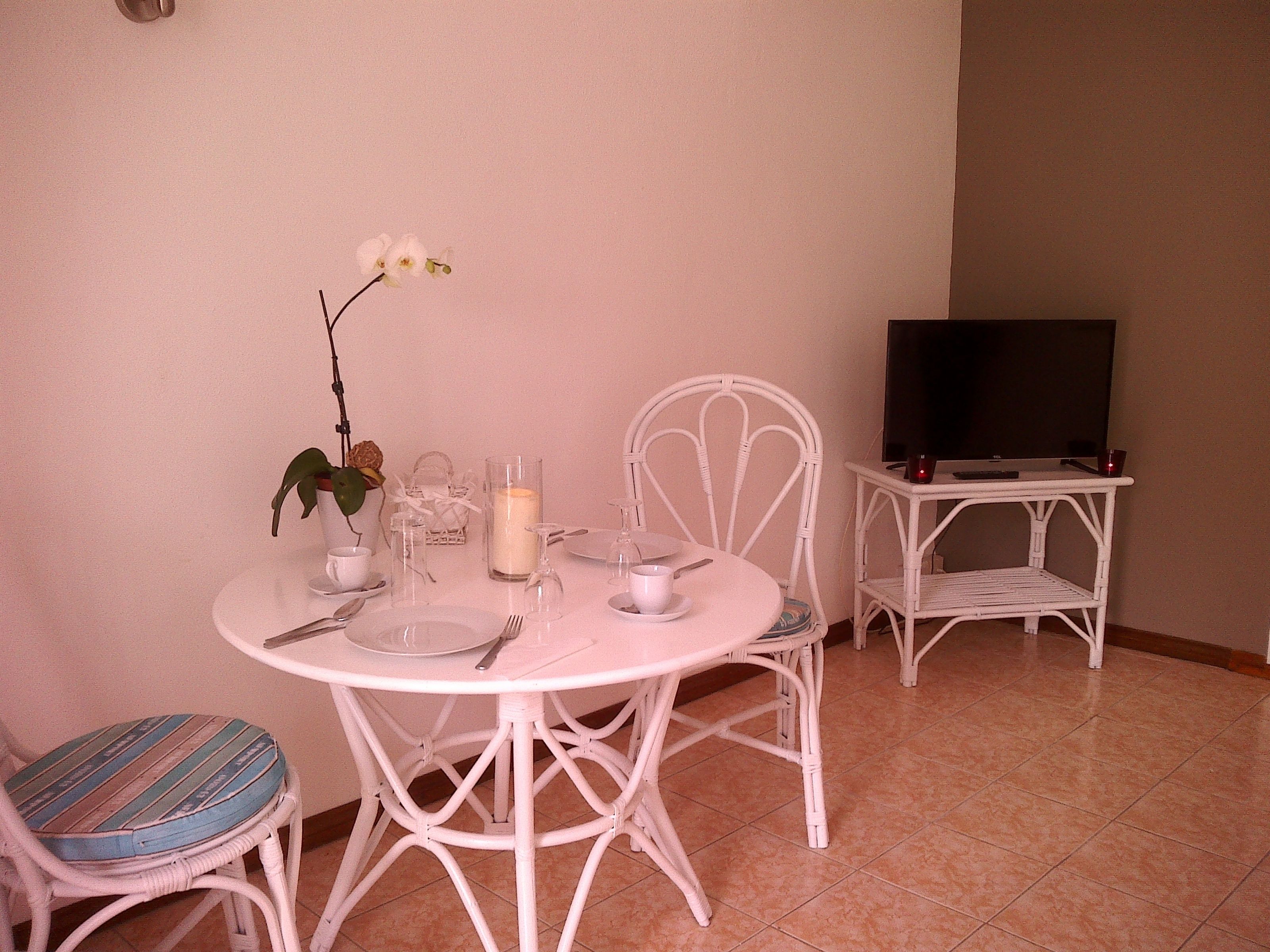 A dining area with a round table set for two, white chairs, a small side table with a candle, and a television on a stand.