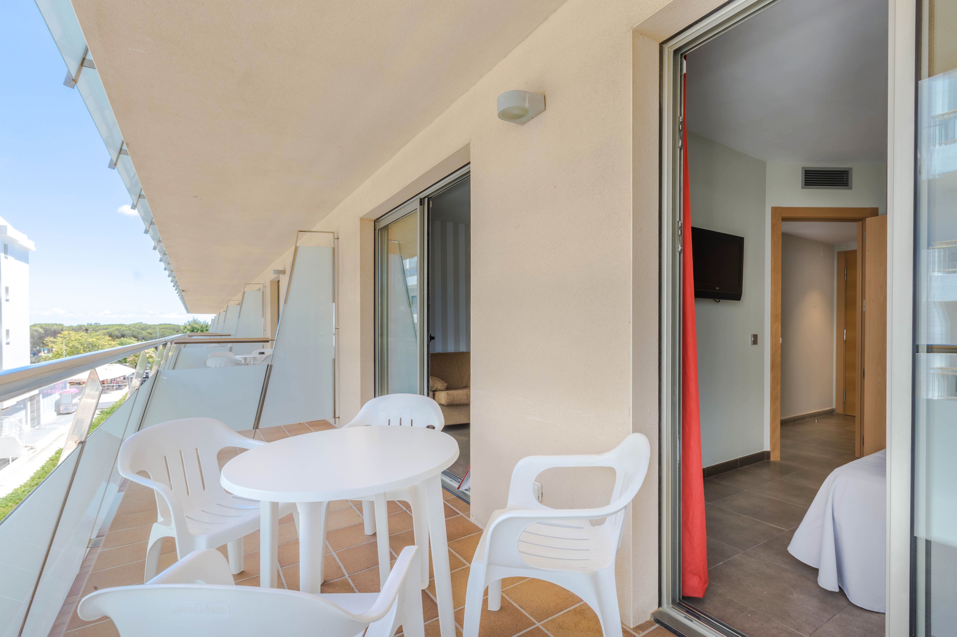 A balcony with white plastic chairs and a table, a sliding glass door leading to a room with a bed and a TV.