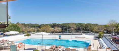 Zona de piscina con sillones de descanso y sombrillas, un trampolín y un cielo azul despejado.