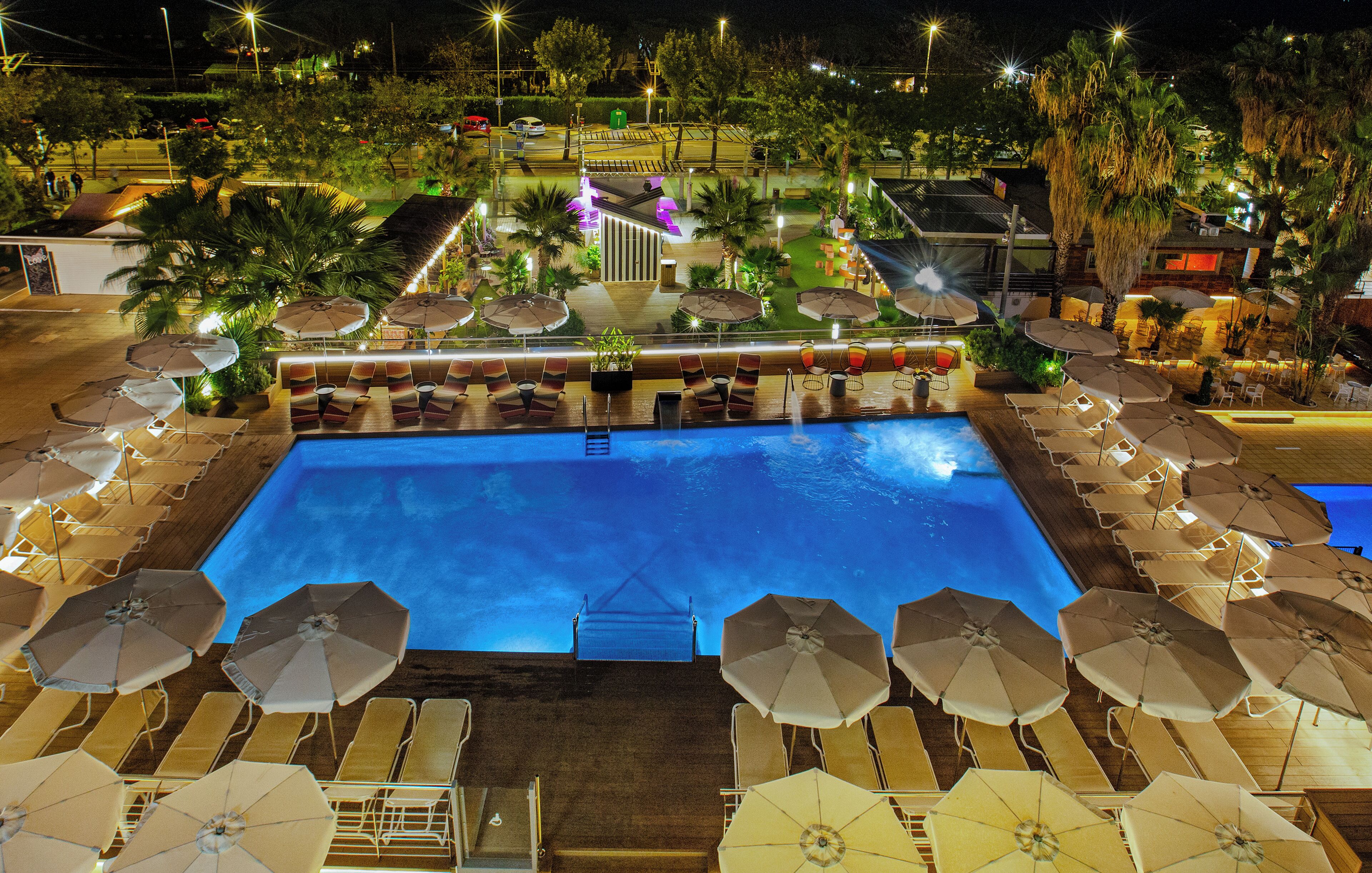 A swimming pool with sun loungers and umbrellas, surrounded by palm trees and a well-lit deck area.