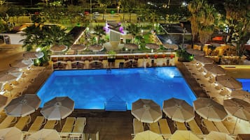 A swimming pool with sun loungers and umbrellas, surrounded by palm trees and a well-lit deck area.