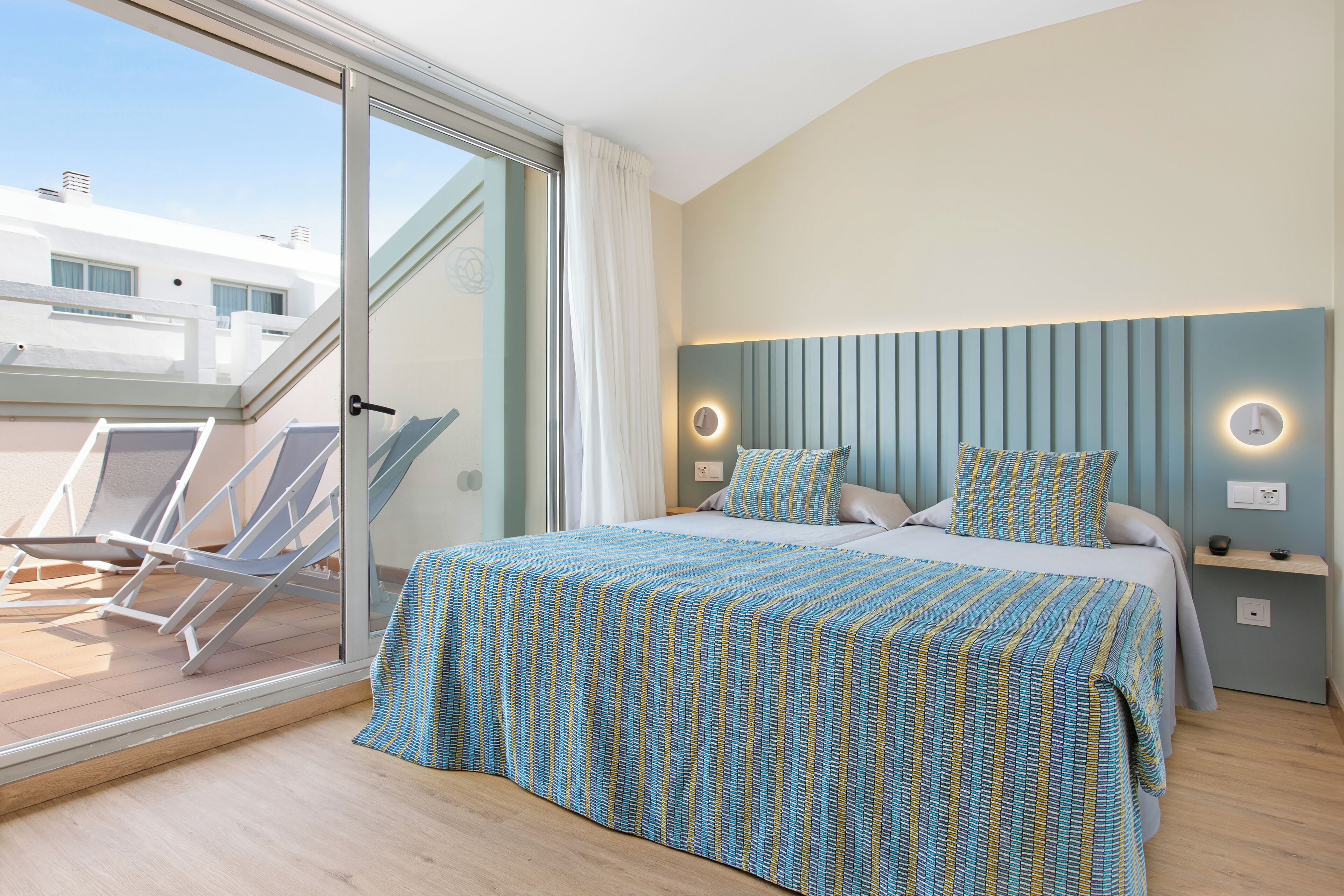 A bedroom with a bed, a nightstand, a wall-mounted light, a window with a curtain, and a balcony with chairs.