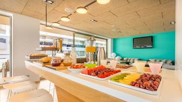 A breakfast bar with a variety of fruits and beverages, white bar stools, and a teal-colored wall.
