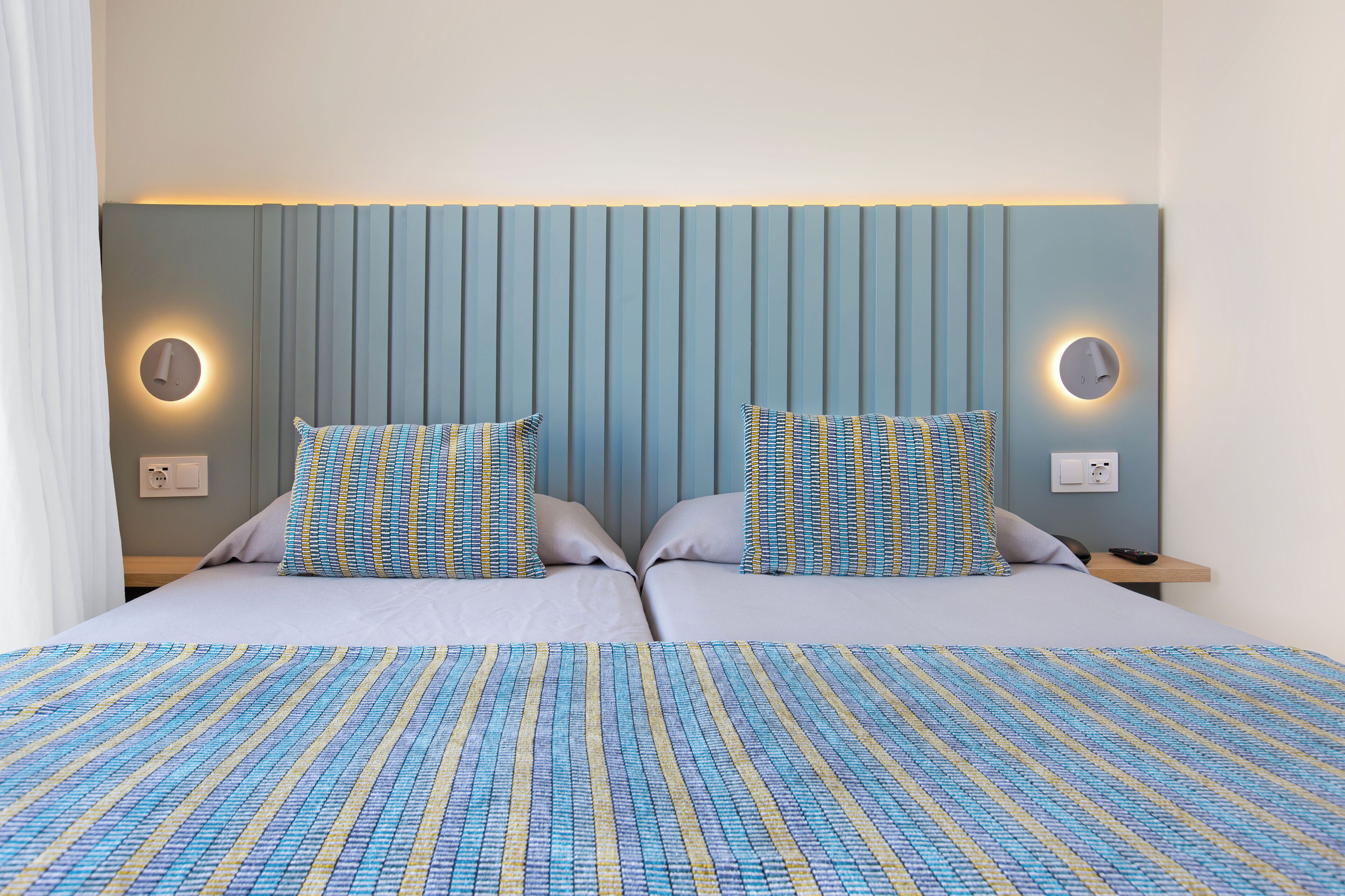 A double bed with a striped bedspread, two matching pillows, a headboard with vertical slats, and wall-mounted reading lights.