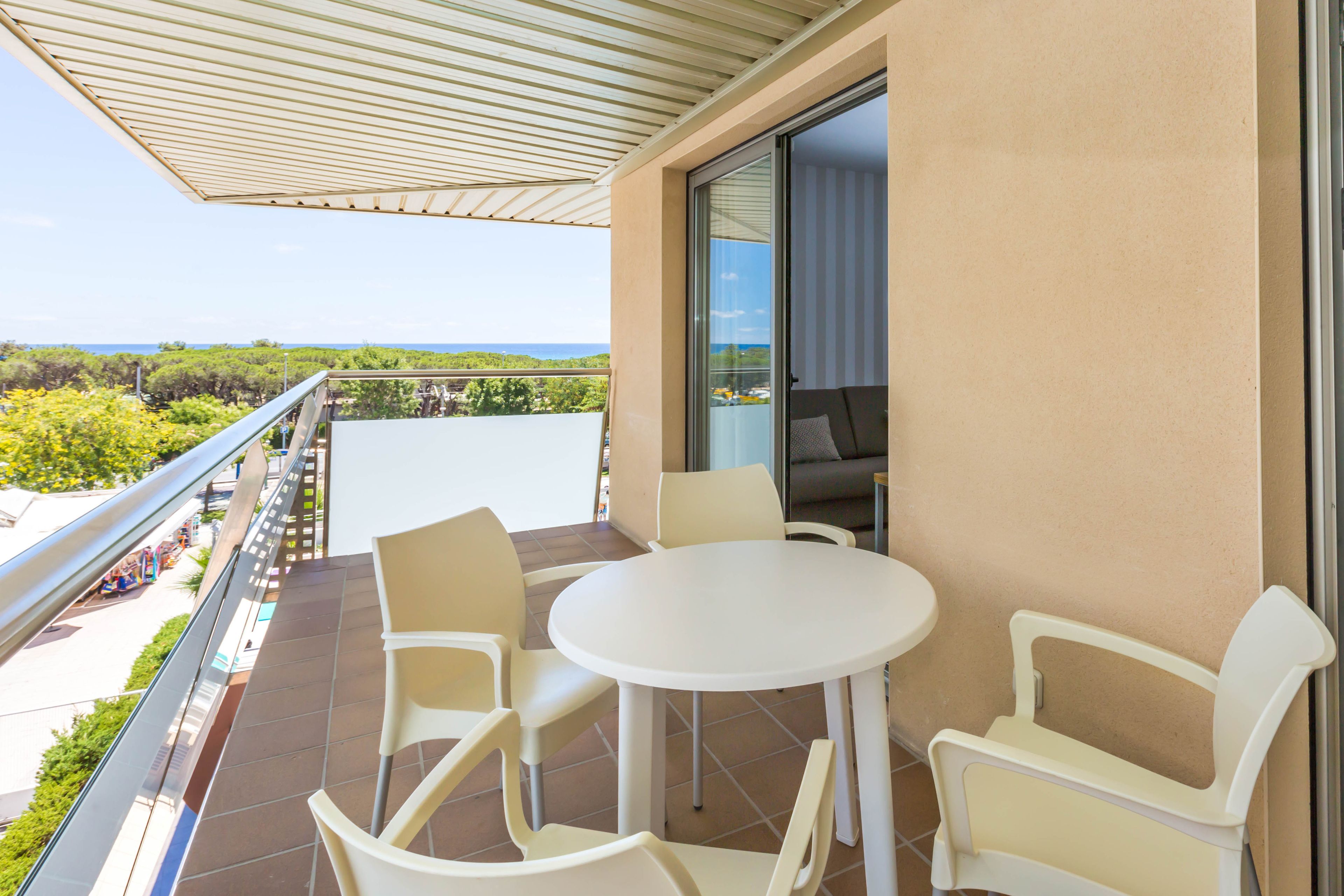 Ein Balkon mit weißem Tisch und Stühlen, einer Glastür und Blick auf das Meer und Grünflächen.