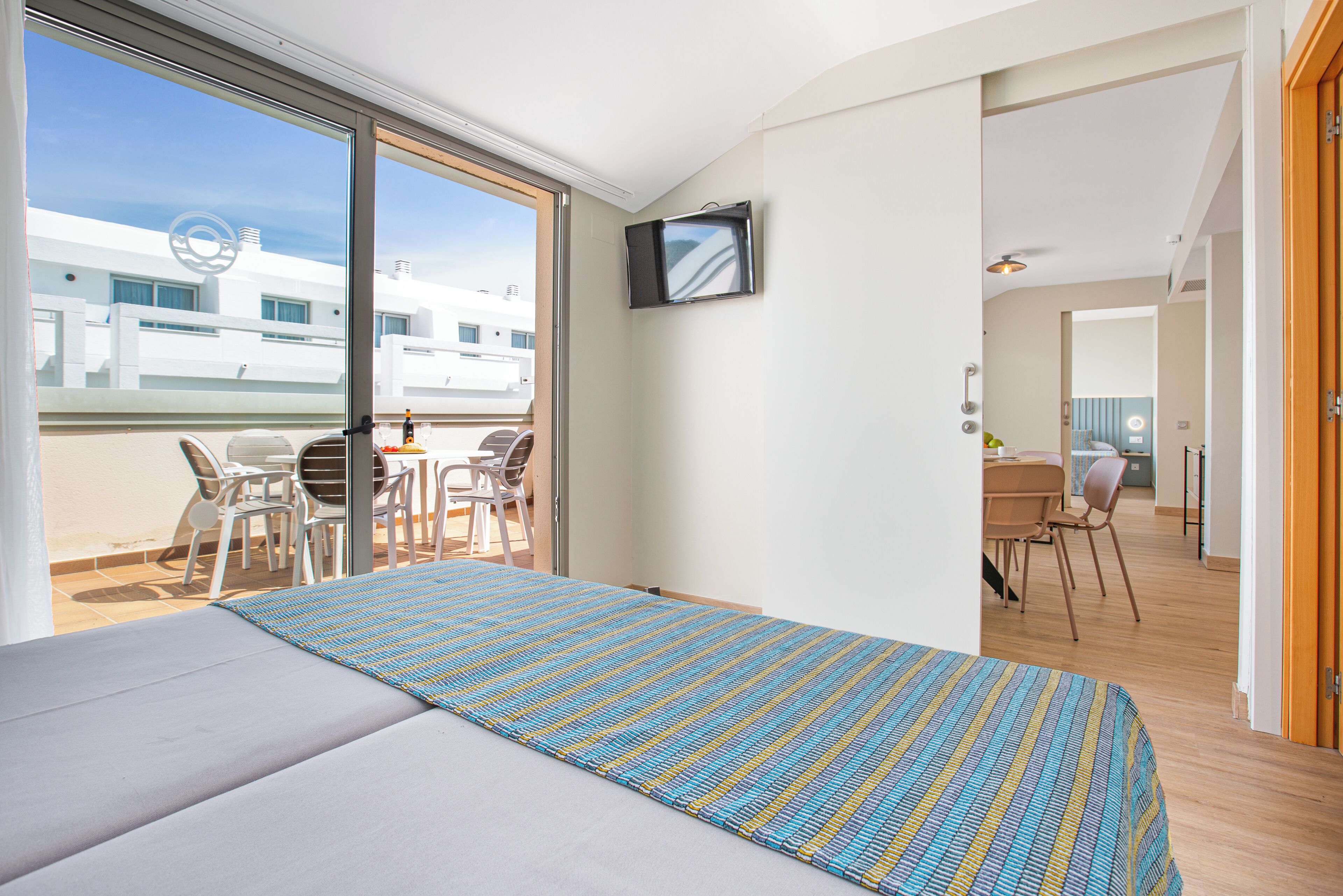 A hotel room with a bed, a TV, a balcony with chairs, and a view of buildings.