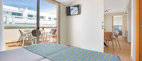 A hotel room with a bed, a TV, a balcony with chairs, and a view of buildings.