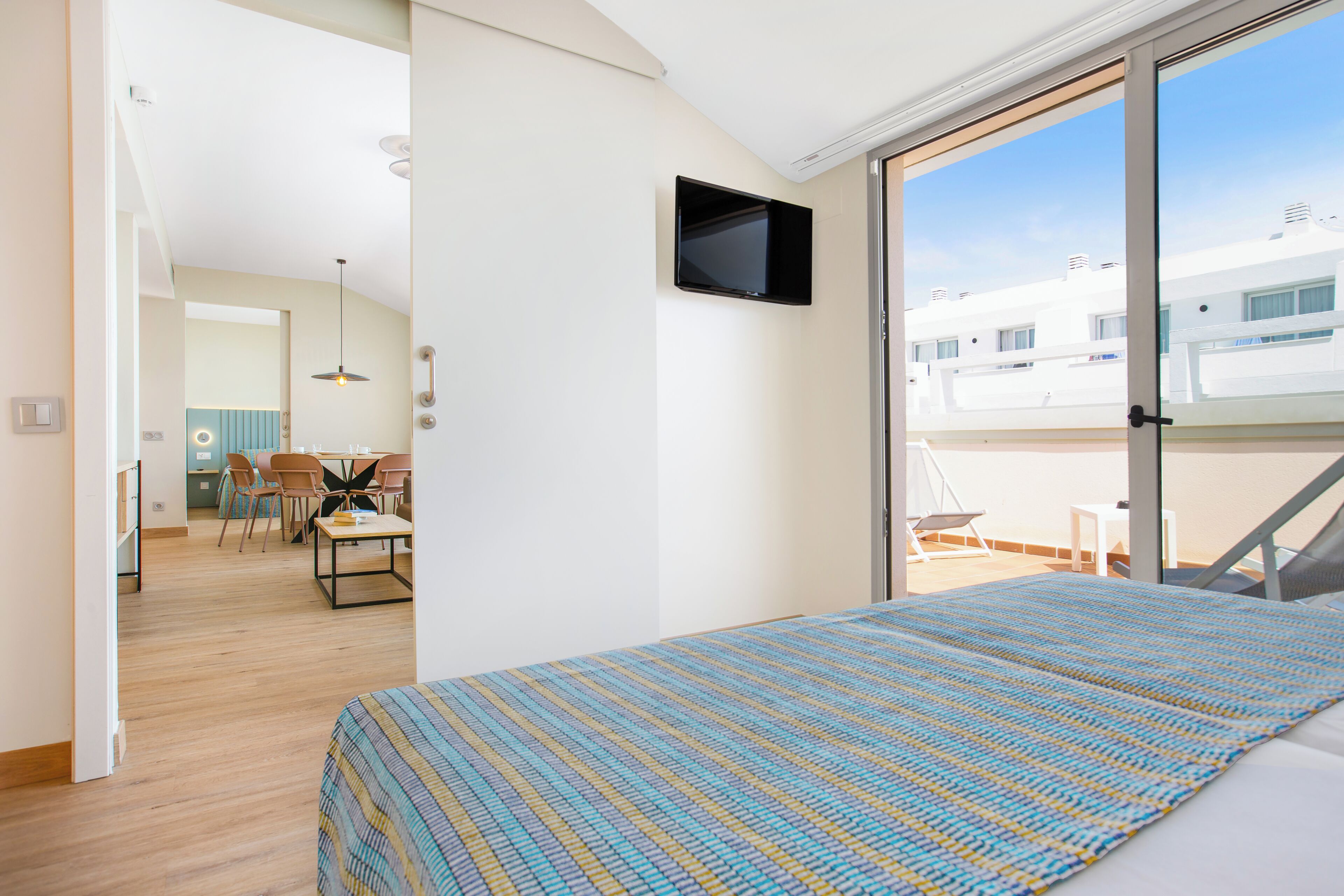A bedroom with a bed, a TV mounted on the wall, and a balcony with a view of buildings.