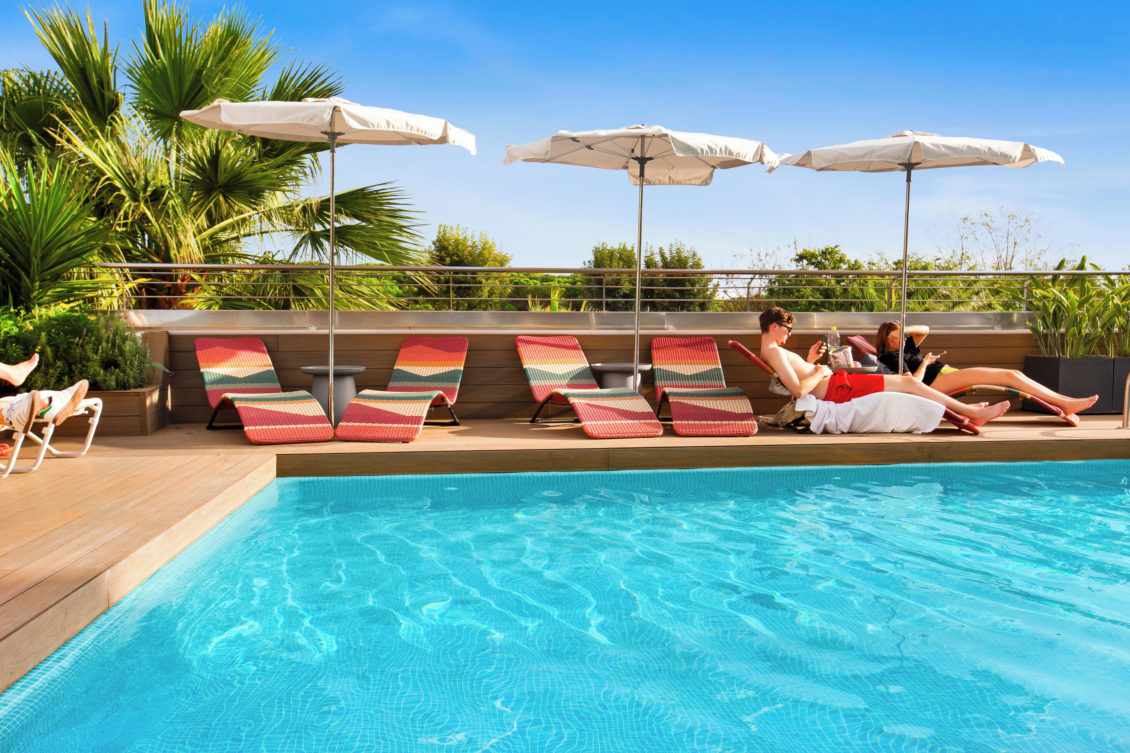 A poolside area with lounge chairs and umbrellas, surrounded by greenery.